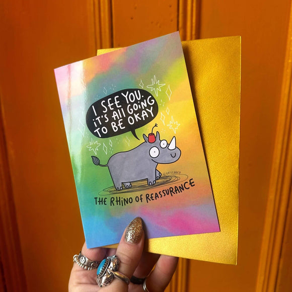 funny A6 greeting card featuring The Rhino of Reassurance designed by Katie Abey. The Rhino is wearing a red hat with a speech bubble coming from it's mouth that reads 'I see you it's all going to be okay'. The card is multicoloured with a gold envelope. 
