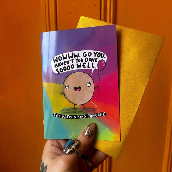 funny congratulations A6 card featuring The Patronising Pancake holding a purple balloon with a smug expression and speech bubble that reads 'wowww. Go you. Haven't you donesoooo well.' on a rainbow coloured card with pairing gold envelope. Designed by Katie Abey.
