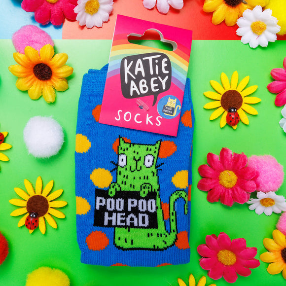 Blue socks with orange and yellow polka dots and green cat illustration holding a black sign that reads 'poo poo head'. There is a pink cardboard label attached to the socks with a rainbow and Katie Abey logo on. The socks are lying on a green background with flowers and ladybugs.