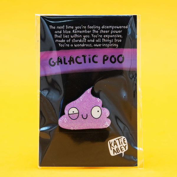 Photo of Katie Abey’s Galactic Poo Glitter Enamel Pin Badge in packaging, with a motivational message above the pin and a bold “Galactic Poo” title. The purple glitter poo character has a goofy, spaced-out expression and is displayed on a black backing card.