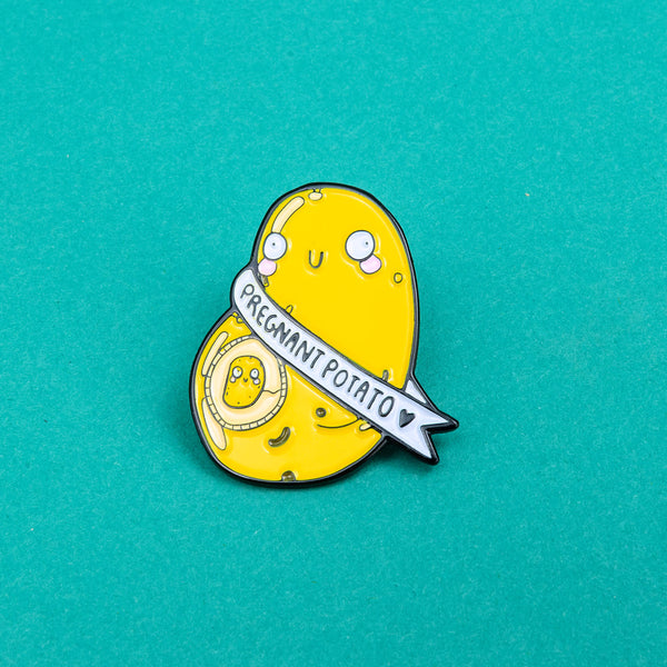enamel pin badge shaped like a potato with a mini potato in its belly space and a banner around it that says pregnant potato. potato illustrated by Katie Abey