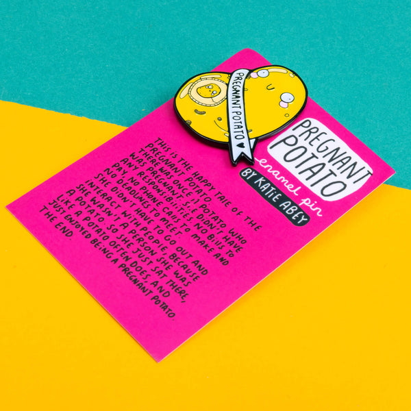 enamel pin badge shaped like a potato with a mini potato in its belly space and a banner around it that says pregnant potato. It is on a pink backing card telling the tale of the potato illustrated by Katie Abey