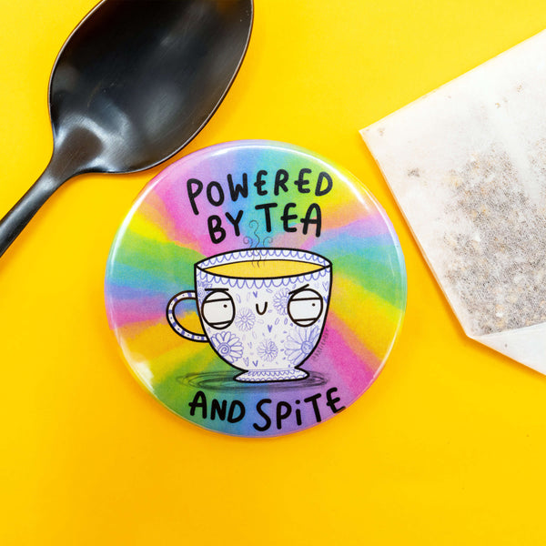 Katie Abey badge with the slogan “Powered by Tea and Spite,” lying on a yellow background next to a black spoon and a used teabag. The badge shows a unimpressed floral teacup character on a rainbow-coloured background.