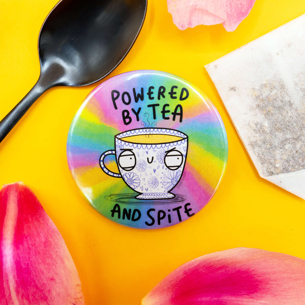 Whimsical badge reading “Powered by Tea and Spite,” placed on a yellow surface with pink flower petals, a teabag, and a black spoon. The design features a sassy floral teacup on a colourful rainbow backdrop.