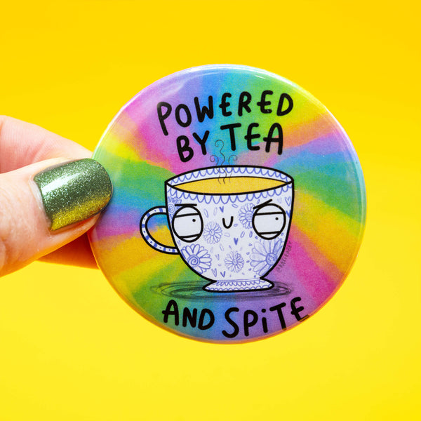Close-up of a hand holding a round Katie Abey badge against a yellow background. The design reads “Powered by Tea and Spite” and shows a moody floral teacup on a vibrant rainbow background. The hand has sparkly green nail polish.