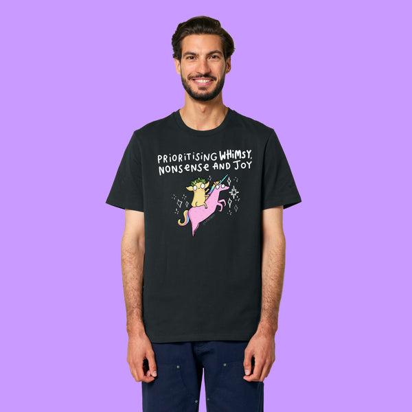 Front view of a black Katie Abey Prioritising Whimsy, Nonsense and Joy adults graphic tee worn by a smiling model. Features a colourful unicorn illustration and playful typography celebrating whimsy, nonsense and joy.