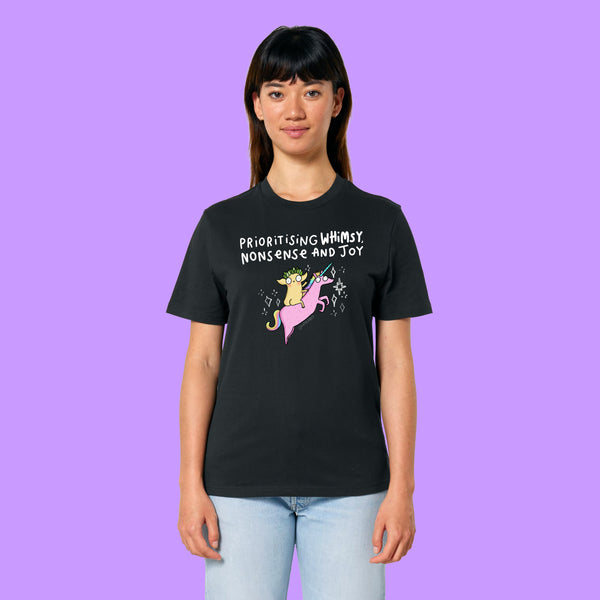 Front view of a black Katie Abey Prioritising Whimsy, Nonsense and Joy adults graphic t-shirt worn by a model. The whimsical illustration shows a quirky character riding a pink unicorn with playful lettering, photographed on a purple background.
