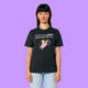 Front view of a black Katie Abey Prioritising Whimsy, Nonsense and Joy adults graphic t-shirt worn by a model. The whimsical illustration shows a quirky character riding a pink unicorn with playful lettering, photographed on a purple background.
