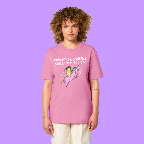 Front view of a Bubble Pink Katie Abey Prioritising Whimsy, Nonsense and Joy adults graphic tee worn by a model with curly hair. The organic cotton unisex t-shirt features a colourful unicorn illustration and playful typography celebrating joy and nonsense.