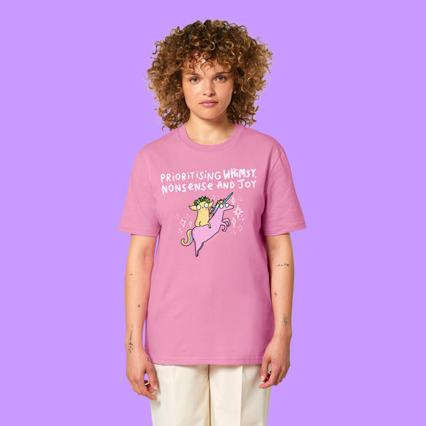 Front view of a Bubble Pink Katie Abey Prioritising Whimsy, Nonsense and Joy adults graphic tee worn by a model with curly hair. The organic cotton unisex t-shirt features a colourful unicorn illustration and playful typography celebrating joy and nonsense.