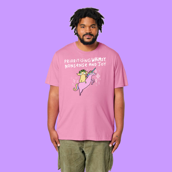 Front view of a Bubble Pink Katie Abey Prioritising Whimsy, Nonsense and Joy adults graphic t-shirt worn by a model. The playful design features a quirky character riding a pink unicorn with whimsical lettering.