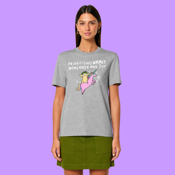 Front view of a heather grey Katie Abey Prioritising Whimsy, Nonsense and Joy adults graphic t-shirt worn by a model. The whimsical illustration shows a character riding a pink unicorn with playful text, styled with a green skirt.