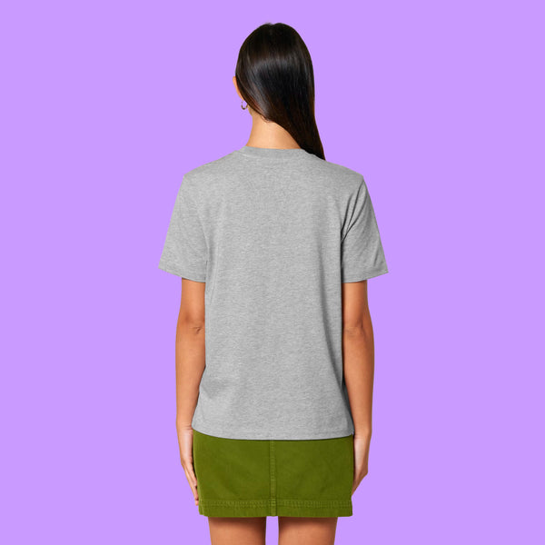 Back view of a heather grey Katie Abey graphic t-shirt worn by a model against a purple background. The organic cotton unisex adults tee shows a relaxed fit and plain back, styled with a green skirt.
