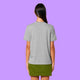 Back view of a heather grey Katie Abey graphic t-shirt worn by a model against a purple background. The organic cotton unisex adults tee shows a relaxed fit and plain back, styled with a green skirt.
