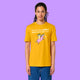 Front view of a Spectra Yellow Katie Abey Prioritising Whimsy, Nonsense and Joy adults graphic tee worn by a model. The design shows a quirky creature riding a pink unicorn with playful text celebrating joy and nonsense.