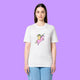 Front view of a white Katie Abey Prioritising Whimsy, Nonsense and Joy adults graphic t-shirt worn by a model against a purple background. The organic cotton unisex tee features a playful illustration of a quirky character riding a pink unicorn with whimsical lettering.