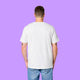 Back view of a white Katie Abey Prioritising Whimsy, Nonsense and Joy adults tee worn by a model with visible arm tattoos. The unisex organic cotton t-shirt shows the plain back and relaxed fit against a purple background.