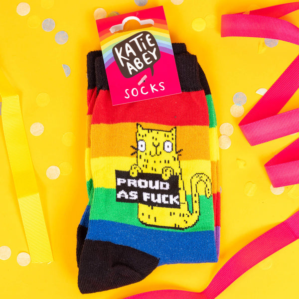 Katie Abey socks with a yellow cat holding a sign saying proud as fuck. They are striped in rainbow colours and lovely and vibrant. The model is stood on a yellow floor with balloons, confetti and ribbons