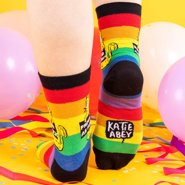A model wearing Katie Abey socks with a yellow cat holding a sign saying proud as fuck. They are striped in rainbow colours and lovely and vibrant. The model is stood on a yellow floor with balloons, confetti and ribbons