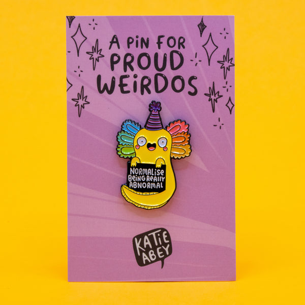 The Normalise Being Really Abnormal axolotl pin badge by Katie Abey shown on a purple Proud Weirdos backing card. The yellow axolotl character has rainbow gills, a party hat, and holds a sign promoting embracing your weird side. A colourful enamel pin for proud weirdos and collectors.