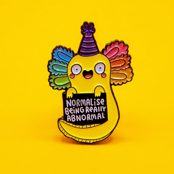 A colourful enamel pin badge by Katie Abey featuring a bright yellow axolotl with rainbow gills and a party hat, holding a sign that says Normalise Being Really Abnormal. Displayed on a vibrant yellow background. A fun, quirky accessory encouraging individuality and embracing your inner weirdo.