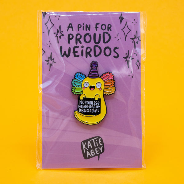 Katie Abey’s Normalise Being Really Abnormal axolotl enamel pin badge displayed on its backing card. The purple card reads A Pin for Proud Weirdos with sparkly doodles. The yellow axolotl character has rainbow gills and a party hat. A cheerful, quirky pin celebrating individuality and weirdness.