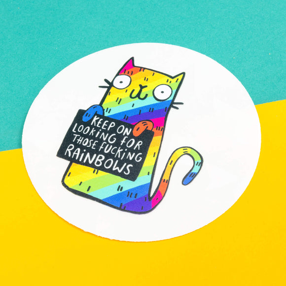 A see through rainbow making sun catching sticker that has a rainbow cat holding a sign saying keep on looking for those fucking rainbows it is laying flat with its backing on