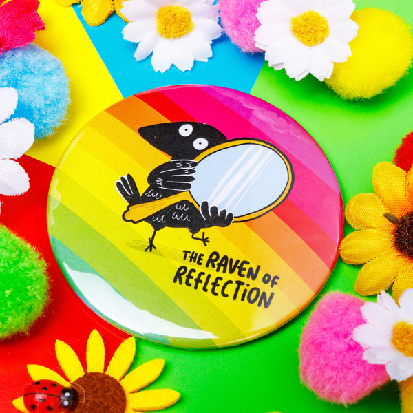 a circular shaped rainbow coloured pocket mirror with the raven of reflection on the front illustrated by Katie Abey