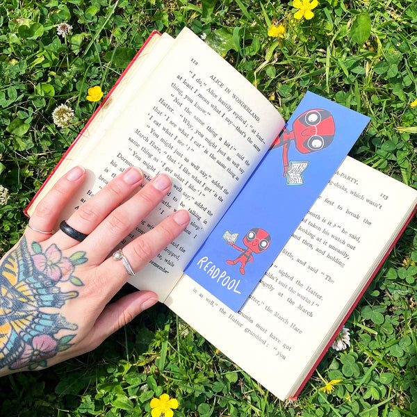 Deadpool Readpool bookmark laying face up in an open book laying on grass. The bookmark is laser printed card with a blue background and two deadpool characters smiling reading books with bottom text reading 'Readpool'. Designed by Katie Abey in the UK and printed with a silk finish so they are wipe clean.