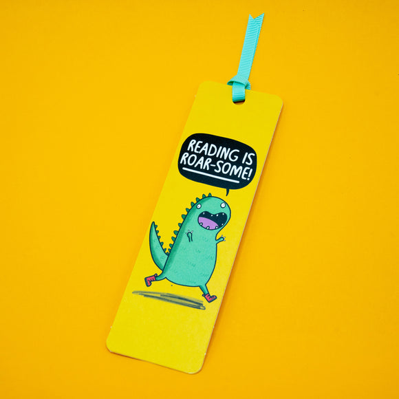 Angled view of Reading is Roar Some Dinosaur Bookmark by Katie Abey featuring cheerful green dinosaur illustration and bold text on yellow background, finished with turquoise ribbon, unique gift for readers and stationery lovers.