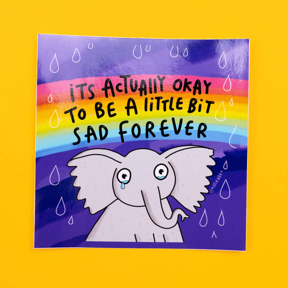 Katie Abey vinyl sticker showcasing a sad-looking cartoon elephant with a tear, set against a purple background with a rainbow above. The sticker reads 'It’s Actually Okay to Be a Little Bit Sad Forever' in comforting black letters, with small raindrop details around the text.