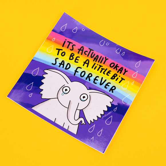 Katie Abey vinyl sticker showcasing a sad-looking cartoon elephant with a tear, set against a purple background with a rainbow above. The sticker reads 'It’s Actually Okay to Be a Little Bit Sad Forever' in comforting black letters, with small raindrop details around the text.