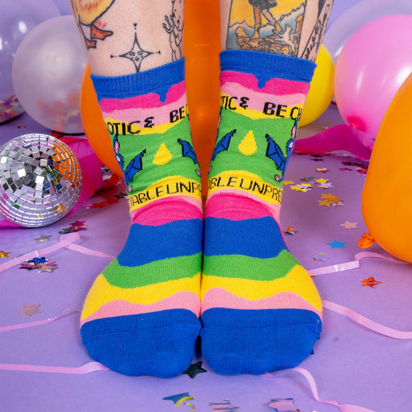 A pair of socks worn by a white model with tattoos. The socks have wavy multi coloured chunks and a drawing by Katie Abey of a bat with text saying be chaotic & Unpredictable