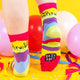 A model wearing Katie Abey socks with yellow, pink, blue, purple and mint green swirls with living vibrantly written on the top. They are striped in rainbow colours and lovely and vibrant. The model is stood on a yellow floor with balloons, confetti and ribbons