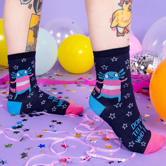 A white tattoo model wearing socks by Katie Abey with Axolotls in trans pride colours and stars with text saying don't tell me who I am on the front. The socks have pink toes and blue heels