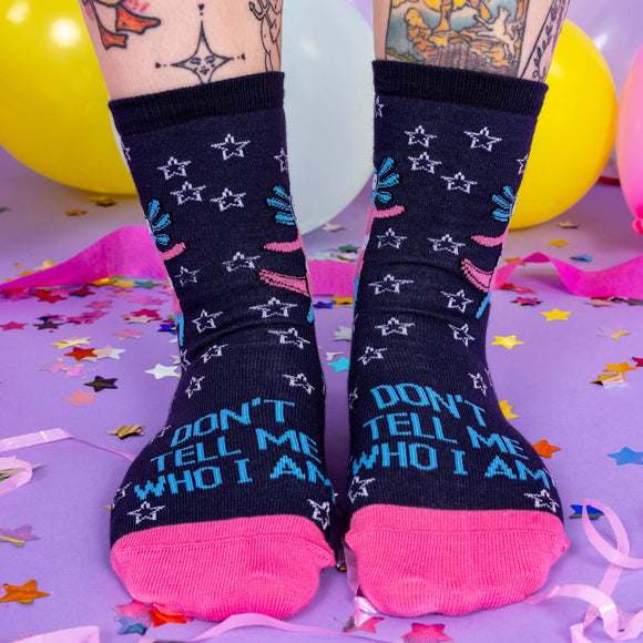 A white tattoo model wearing socks by Katie Abey with Axolotls in trans pride colours and stars with text saying don't tell me who I am on the front. The socks have pink toes and blue heels