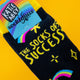 Close-up of Katie Abey’s “The Socks of Success” socks on a yellow background. The black socks feature yellow text, rainbow shooting stars, and playful motifs, with the Katie Abey Socktopus label visible. A quirky, positive design from the UK illustrator known for celebrating weirdness and joy.