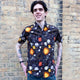 Jake a man with tattoos on his arms and dark hair is wearing Run & Fly x Katie Abey Solar Witch Short Sleeve Shirt paired with blue jeans. The shirt is black with an all over print of Katie Abey witchy illustrations. Jake is smiling at looking into the distance with his hands on his hips in front of a brick wall outside in Hove.