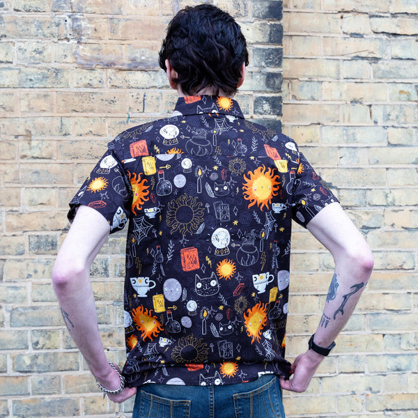 Jake a man with tattoos on his arms and dark hair is wearing Run & Fly x Katie Abey Solar Witch Short Sleeve Shirt paired with blue jeans. The shirt is black with an all over print of Katie Abey witchy illustrations. Jake is stood with his back to the camera in front of a brick wall and is pulling the shirt down at the back.
