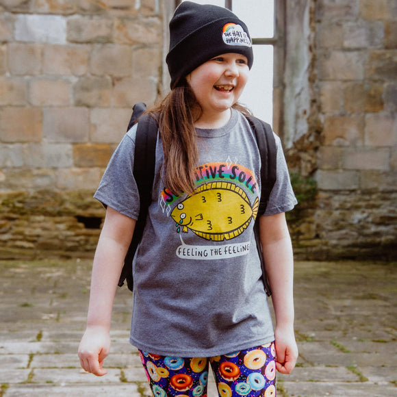 The sensitive sole t shirt worn by a chid smiling outside. The grey t shirt has an illustration of a yellow sole with tears from it's eyes with a rainbow above it with the text sensitive sole written in front. There is a speech bubble from the sole's mouth that reads feeling the feelings