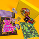 A selection of Katie Abey's galactic-themed items, including a "Give Me Some Space" poster, a glittery poop sticker, and an alien-patterned cloth. The bright, bold colours and fun designs celebrate individuality and creativity.