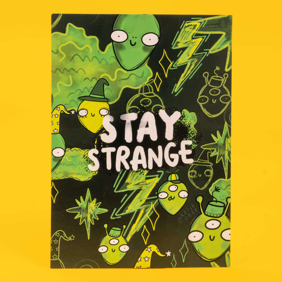 Colourful postcard by Katie Abey featuring the phrase "Stay Strange" in bold white letters. The background is filled with quirky, green alien characters and lightning bolts on a black background. Some aliens wear hats and have various expressions. The design is playful and encourages embracing individuality. The artist's handle, @katieabey, is subtly included. The background is bright yellow