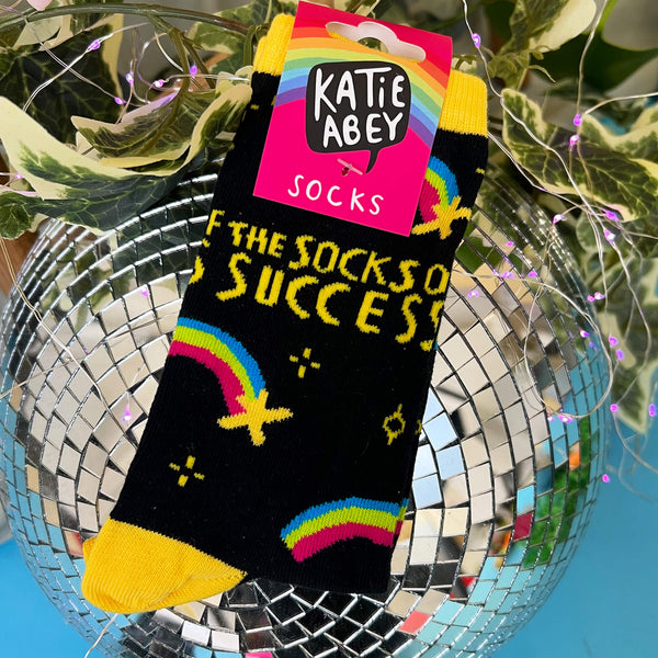 The socks of success, designed by Katie Abey in the UK, are a black colour with yellow heels, toes and top cuffs with various yellow sparkles, rainbow shooting stars and yellow text reading 'the socks of success'.