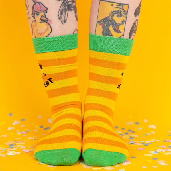 A front-facing shot of the yellow and orange striped socks with green accents, worn on legs with tattoos visible, standing on a confetti-covered floor against a yellow backdrop.