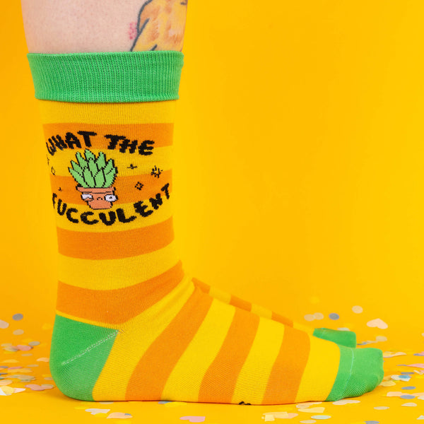 A side view of bright yellow and orange striped socks with green cuffs and toes, featuring a cheeky succulent pot design and the phrase “What the Fucculent?” against a vibrant yellow background sprinkled with confetti.