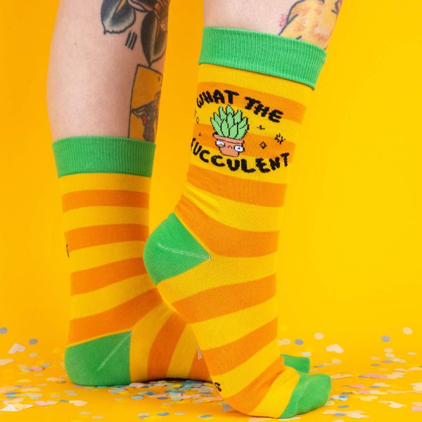 A playful pose showing both feet in the yellow striped socks, highlighting the succulent design on the front and the contrasting green heel and toes, with confetti scattered below.