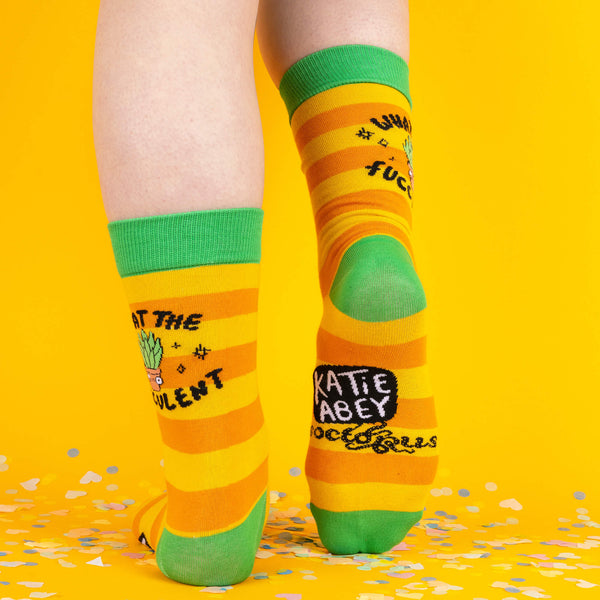 A back view of the striped socks showing “Katie Abey” branding on the sole of one sock, with the “What the Fucculent?” design on the other, all against a bright yellow background with scattered confetti.