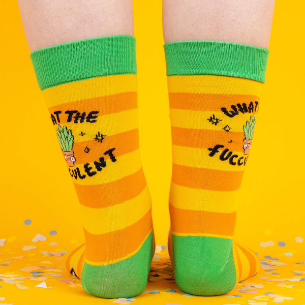  A rear shot of the yellow and orange striped socks with “What the Fucculent?” designs visible on both sides, worn on legs standing against a confetti-strewn yellow floor.