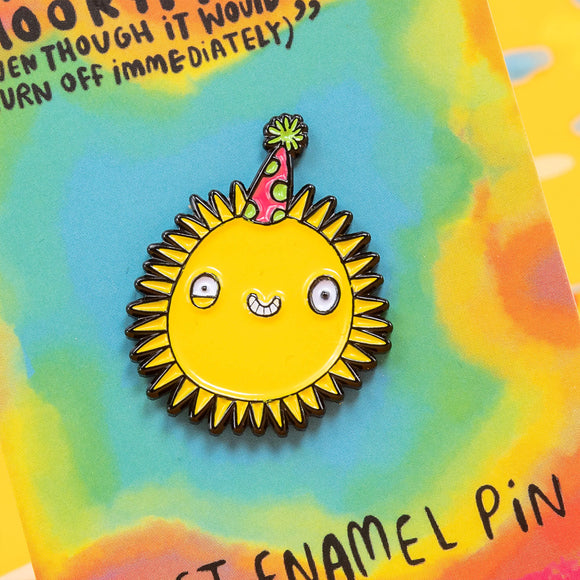 Close up of A soft enamel pin designed by Katie Abey with a sun smiling wearing a party hat it is on a backing card saying the sun has got his hat on hip hip hip hooray (even though it would definitely burn off immediately) 