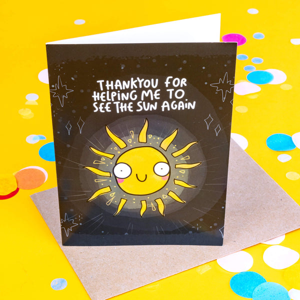 A black greeting card with white twinkling stars and white writing on the front that reads 'thankyou for helping me to see the sun again' and an illustration of a yellow sun with pink cheeks and a smiley face